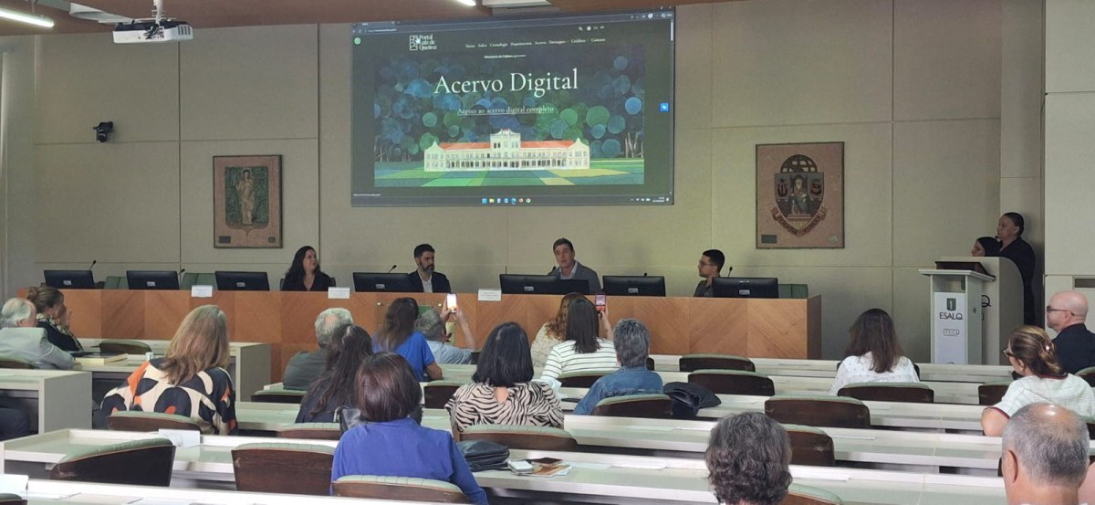 Vis&atilde;o geral de um audit&oacute;rio lotado durante evento do Portal Luiz de Queiroz. Ao fundo, em uma mesa de honra, quatro debatedores est&atilde;o sentados. Acima deles, uma tela projeta a p&aacute;gina inicial do site "Acervo Digital" com a ilustra&ccedil;&atilde;o do pr&eacute;dio hist&oacute;rico da ESALQ. Na plateia, v&aacute;rias pessoas assistem &agrave; apresenta&ccedil;&atilde;o.
